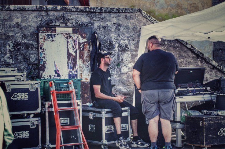 Due tecnici al lavoro, seduti su scatole di attrezzatura audio e video, discutono mentre una tenda è montata sullo sfondo.