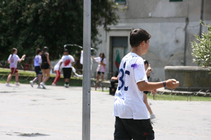 Bambini che giocano all'aperto in una piazza, indossando magliette sporche di vernice, alcuni si divertono mentre altri sono in movimento.