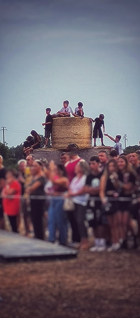 Un gruppo di ragazzi in cima a un grande rotolo di fieno, mentre una folla osserva da vicino.